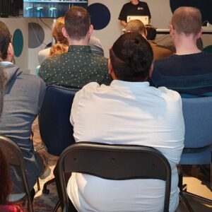 A group of 20 people sitting and looking towards a speaker at a Meetup in a Office-setting.
