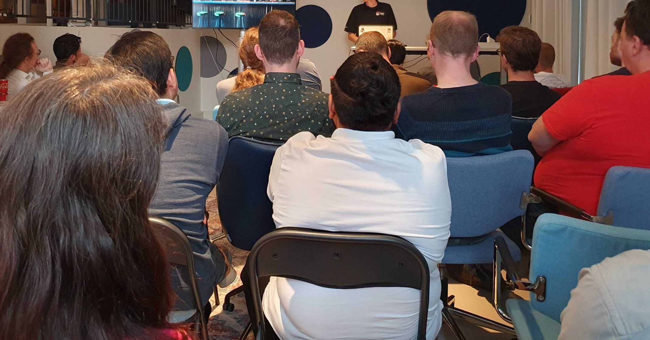 A group of 20 people sitting and looking towards a speaker at a Meetup in a Office-setting.